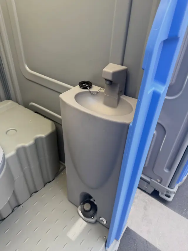 Inside a portable toilet with a small foot-pump sink beside the seat
